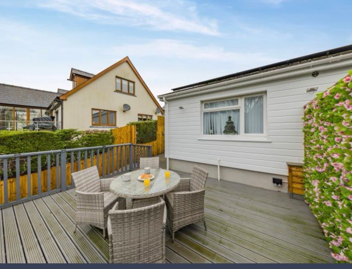 Spacious decked terrace with comfortable outdoor seating, featuring flowering climbing plants and views towards the main cottage and woodland surroundings.
