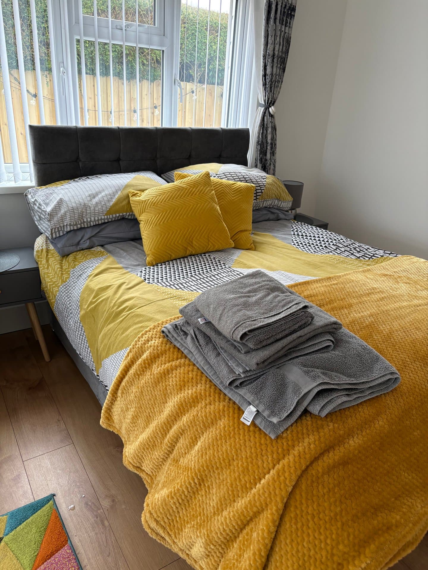 Cosy bedroom with stylish yellow and grey bedding, comfortable black upholstered headboard, and bright bay windows with woodland views. Perfect for a peaceful retreat.